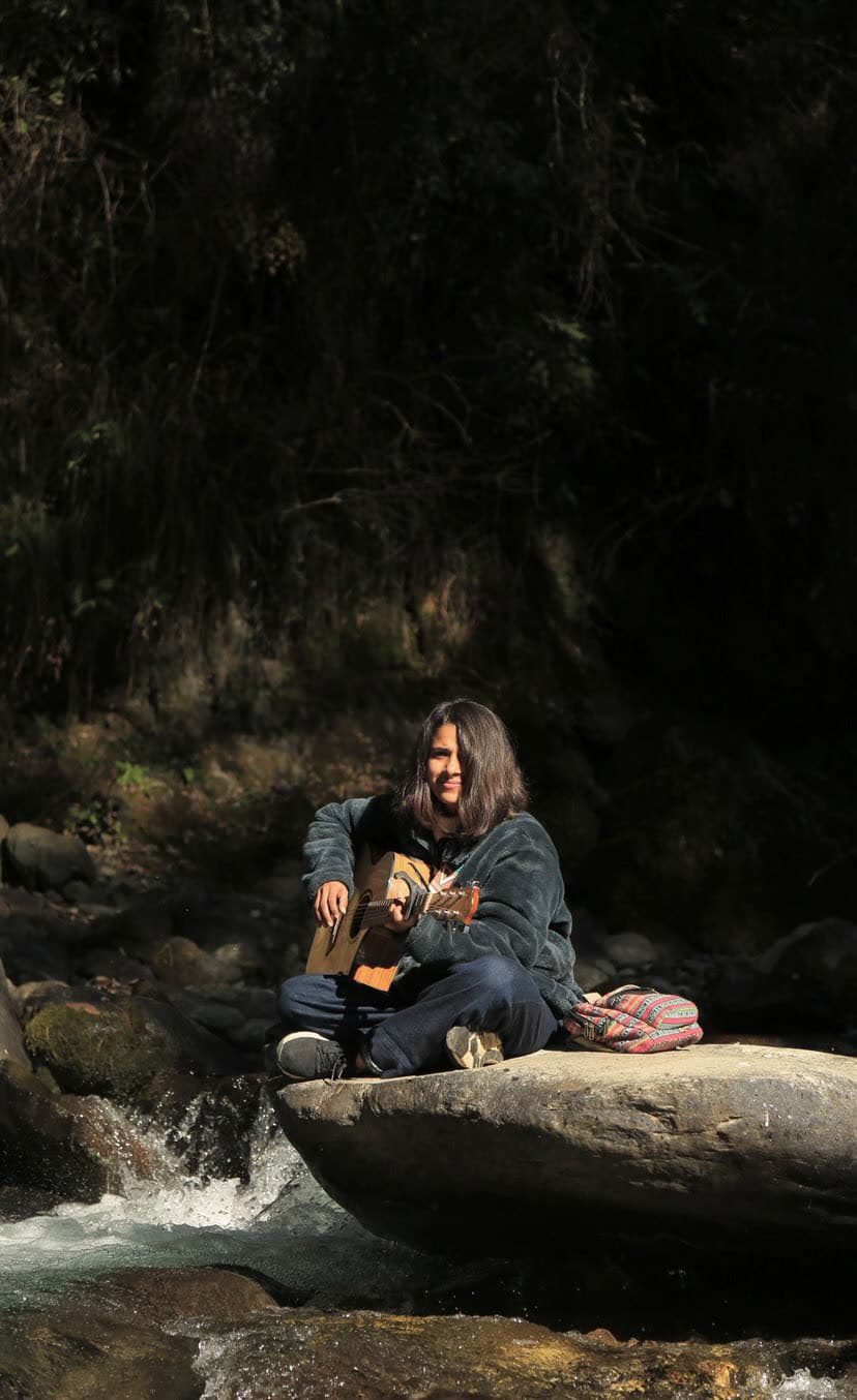 Aashreya playing guitar on a boulder beside a mountain stream